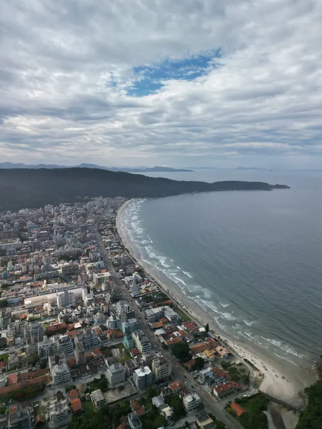 Vista aérea da Praia de Bombas, uma longa faixa de areia curvando entre a cidade e um costão verde, com uma ilha no horizonte sob céu nublado.