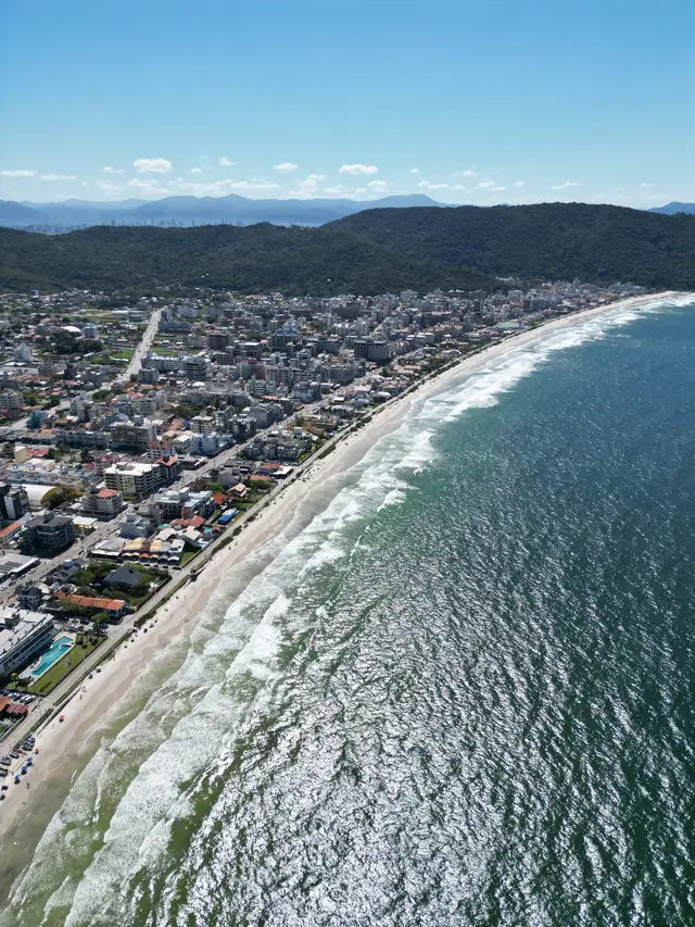 Vista aérea da Praia de Bombas em dia de sol, com muitas ondas brancas na areia e a cidade acompanhando toda a extensão da praia.