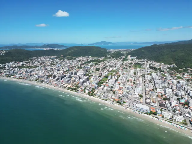Vista aérea ampla da Praia de Bombas e da cidade em dia de sol, encaixadas entre o mar e os morros, com ilhas no horizonte.