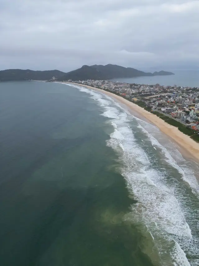 Vista aérea da Praia de Canto Grande pelo lado do Mar de Fora em dia nublado, com ondas quebrando ao longo de uma faixa de areia extensa e a cidade à direita.