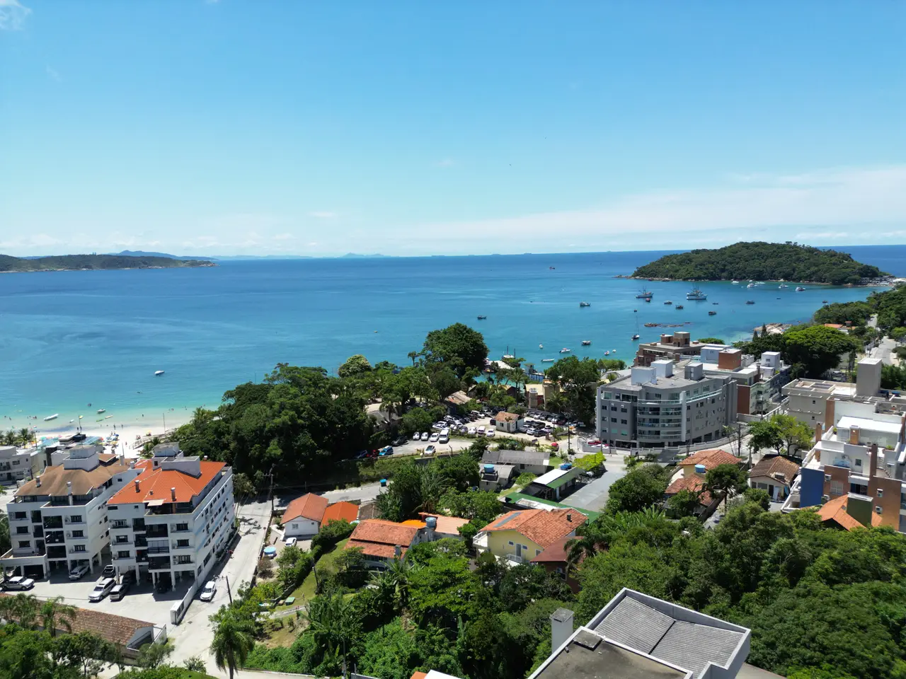 Vista aérea de uma enseada de água azul turquesa em Bombinhas, com faixa de areia e banhistas à esquerda, barcos ancorados e um costão coberto de mata à direita.
