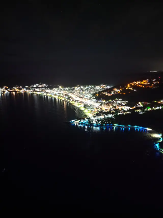 Vista aérea noturna de Bombinhas, com as luzes da cidade acompanhando a curva da praia e refletindo no mar escuro, e uma passarela iluminada sobre a água.