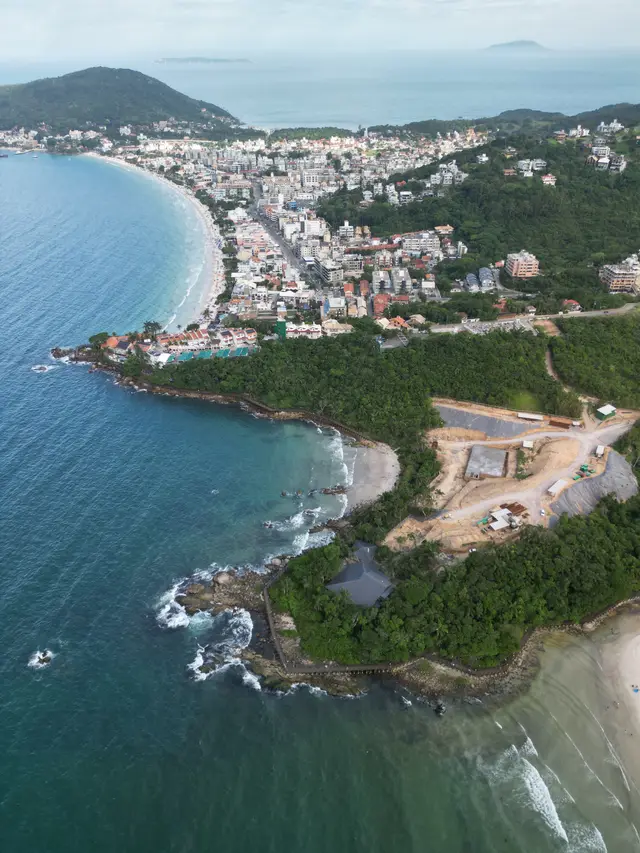 Vista aérea do Centro de Bombinhas com a praia à esquerda e um costão de pedras com pequenas enseadas à direita, e ilhas no horizonte.